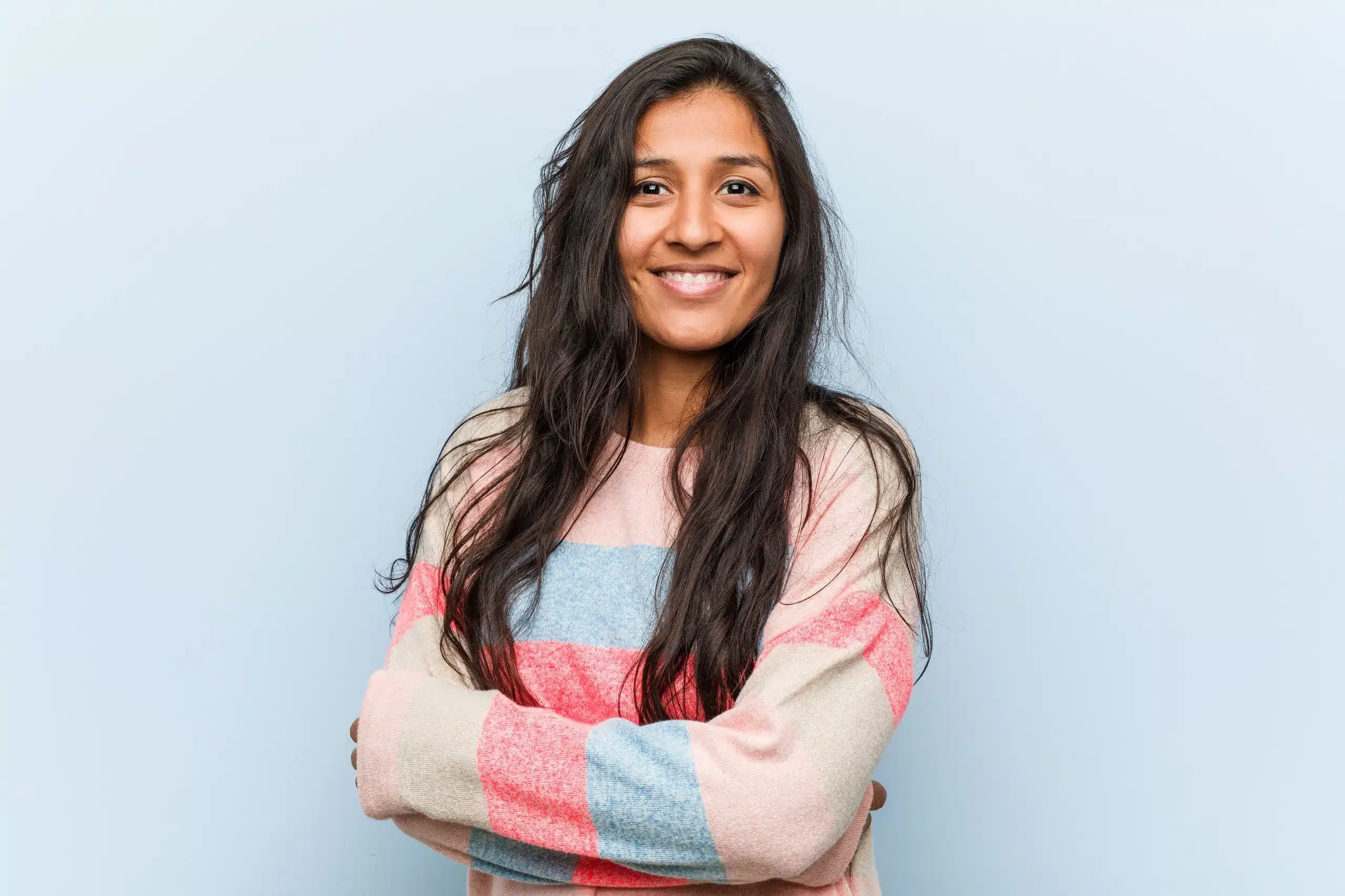 Young woman with long dark hair smiling confidently while standing with arms crossed against a light blue background, wearing a colorful striped sweater for Seashore MD in Little Silver, NJ.
