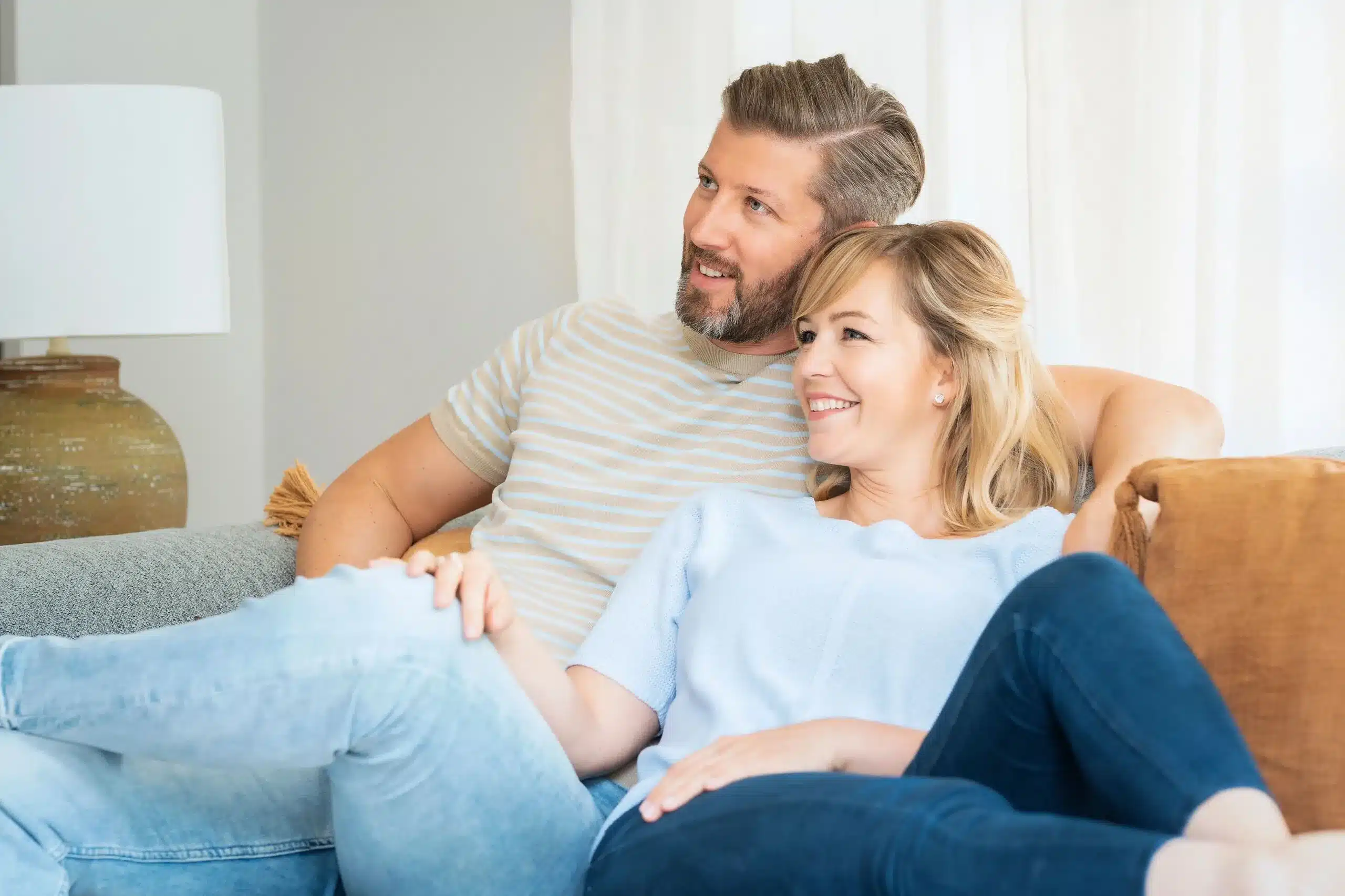 Happy middle-aged couple relaxing together on a cozy couch at home, smiling and enjoying quality time, symbolizing renewed vitality and balance often experienced through Hormone Replacement Therapy at Seashore MD in Little Silver, NJ.