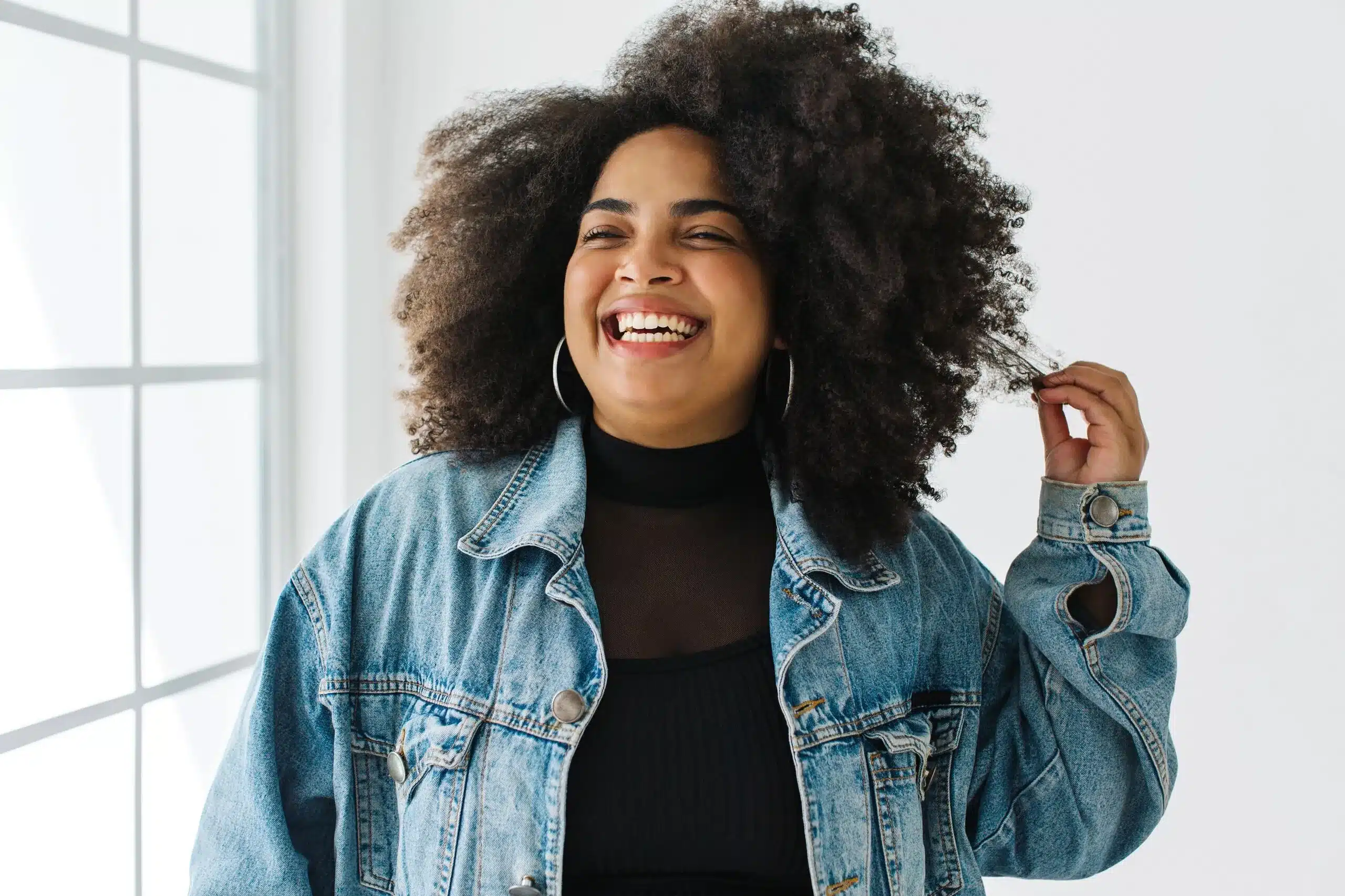 Confident young woman smiling brightly in natural light while touching her curly hair, representing strength and positivity in managing PCOS & hormonal disorders at Seashore MD in Little Silver, NJ.