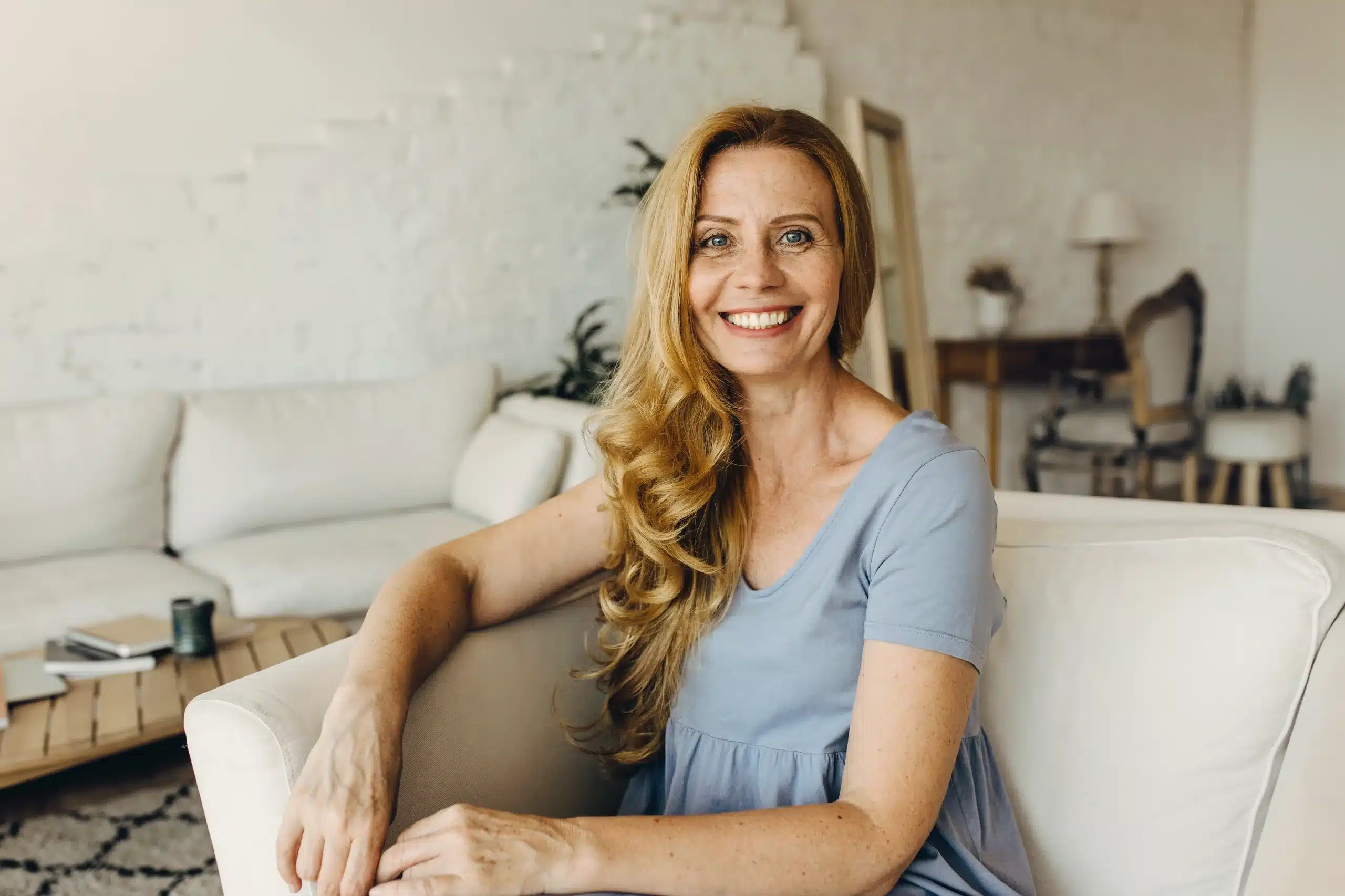 Smiling middle-aged woman with long blonde hair sitting comfortably on a beige couch in a cozy, well-lit living room with a relaxed and welcoming atmosphere at Seashore MD in Little Silver, NJ.