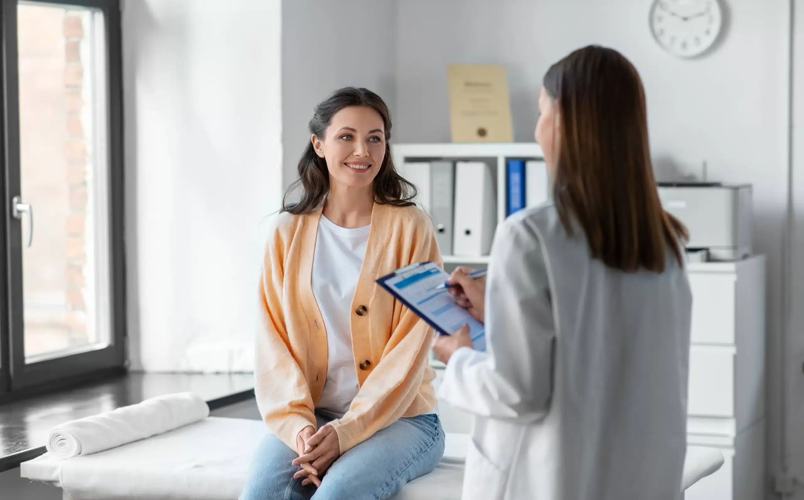 Smiling woman sitting on a medical exam table during a consultation with a healthcare provider—representing Annual Women’s Wellness Exams and preventive health care at Seashore MD in Little Silver, NJ.